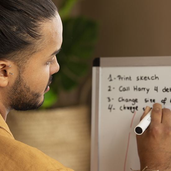 A person with a bun hairstyle and ear piercings is writing on a whiteboard. The whiteboard lists several tasks, including "Print sketch," "Call Harry 4 details," "Change size of," and "Change." A green plant is visible in the background.