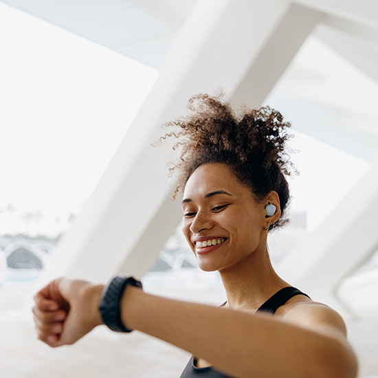 A woman with curly hair, wearing a sleeveless athletic top and earbuds, smiles while looking at her smartwatch. The background features a large, bright architectural structure with geometric lines.