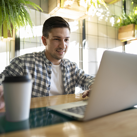 A man in a checkered shirt is smiling and working on a laptop in a bright room with lots of natural light. A to-go coffee cup is placed on the desk beside him. The background features hanging plants and large windows.