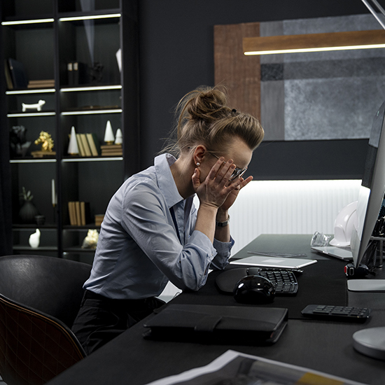 A person sitting at a desk in a modern office with their head in their hands, appearing stressed. The desk has a computer, keyboard, mouse, and a notebook. In the background, there's a black bookshelf with books and decorative items.