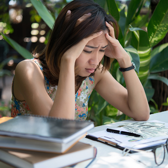 A woman sits at a table outdoors, holding her head in her hands and appearing stressed. She is surrounded by books, notebooks, and papers. Green plants and a soft natural background fill the scene.