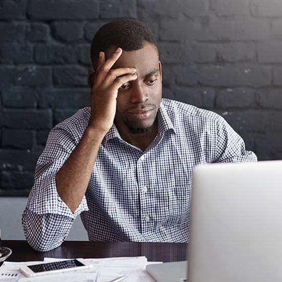 A man wearing a checked shirt sits at a desk, looking at a laptop screen with a pensive expression. He rests his forehead on one hand. The desk has scattered papers and a phone on it. The background features a dark brick wall.