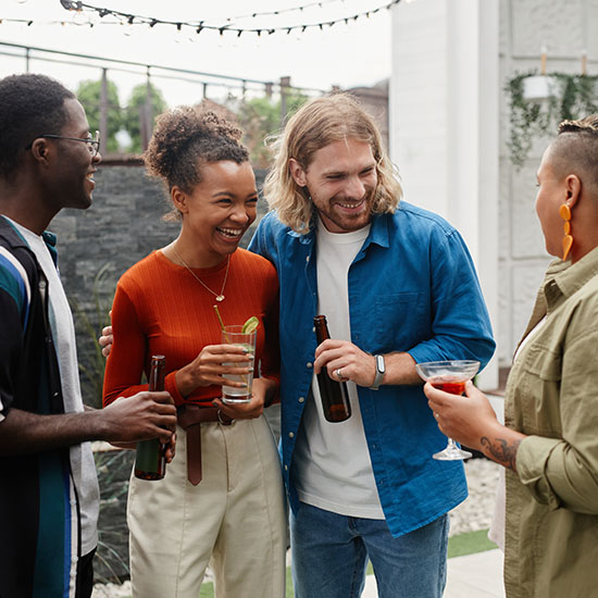 A group of four diverse friends stand outdoors, laughing and talking while holding drinks. Two of them hold beer bottles, one holds a cocktail, and another holds a drink with a straw. They appear to be enjoying a casual, friendly gathering.