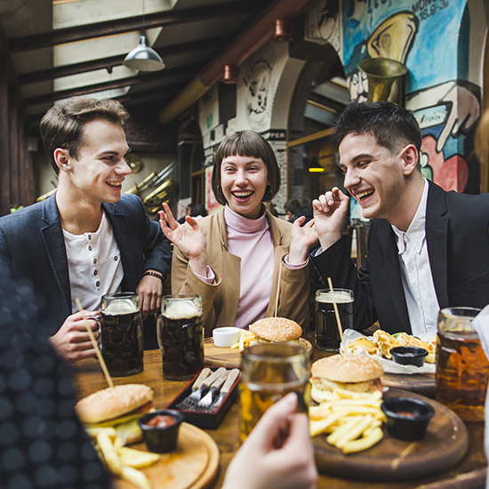 Three young adults are seated at a restaurant table, enjoying burgers, fries, and drinks. They are laughing and appear to be in a lively conversation. The background features artistic murals, adding to the vibrant atmosphere of the restaurant.