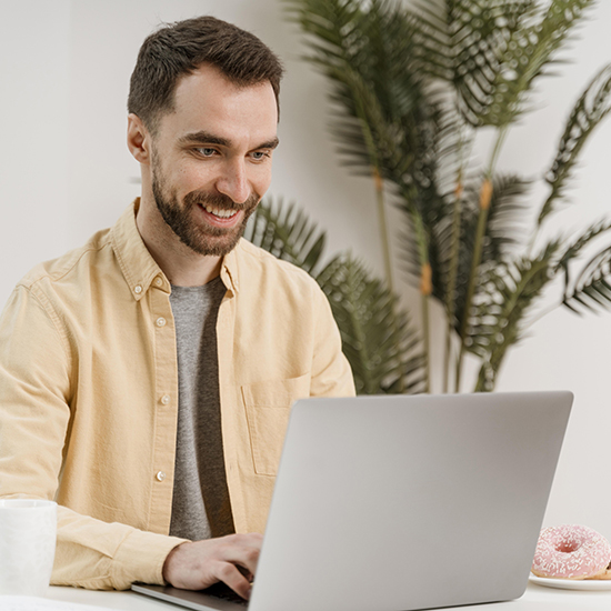 A man with a beard sits at a desk, smiling while looking at a laptop screen. He is wearing a beige shirt over a gray t-shirt. There is a cup and a donut on the desk beside him. In the background, there are green plants.