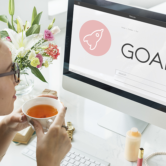 A person holding a cup of tea looks at a computer screen displaying the word "GOALS" with a rocket icon, reflecting on goal setting. A vase of flowers is nearby, and various items are on the desk, including glasses and a watch.