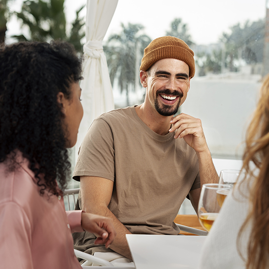 A bearded man, known as a great conversationalist, wearing a brown beanie and beige t-shirt, laughs while sitting at a table with two people. They're outdoors with drinks on the table, and trees visible in the background.