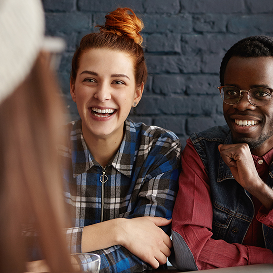 Two people are sitting at a table, smiling as if under a spell. One, with hair in a bun and a plaid shirt, listens intently. The other, a natural conversationalist in glasses and a denim vest, gestures animatedly. They chat indoors against the backdrop of a dark brick wall.