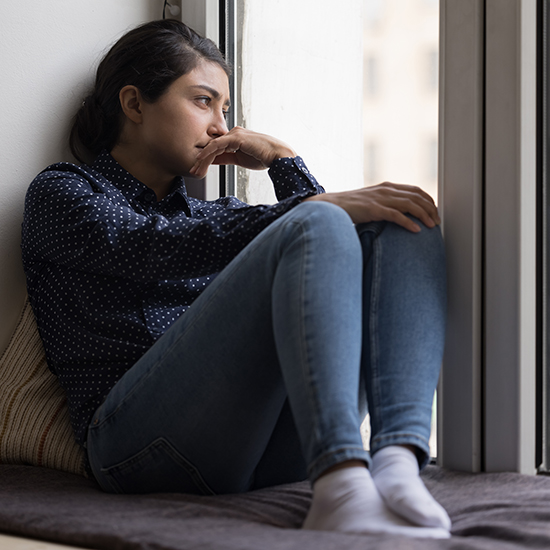 A pensive person sits by a window, resting their chin on their hand. They are wearing a dark polka dot shirt and light blue jeans, looking outside with a contemplative expression that hints at loneliness. Soft light illuminates the scene, evoking the tranquility of self-reflection through hypnotherapy.