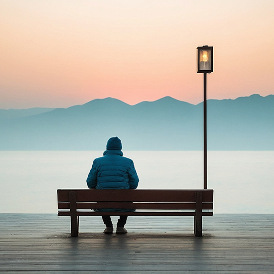 A person in a blue jacket sits on a bench facing a calm body of water at dawn, enveloped by the gentle embrace of loneliness. A lone streetlamp stands nearby. The horizon features a pastel sky with silhouetted mountains in the distance, like nature's own form of hypnotherapy.