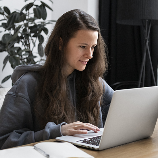 A woman with long brown hair, wearing a gray hoodie, is sitting at a wooden table, typing on her laptop about hypnotherapy. A notebook and pen are in the foreground, while the serenity of a large potted plant fills the background.