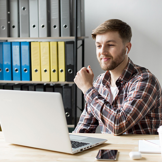 A man with a beard, wearing a plaid shirt and wireless earbuds, sits at a desk using a laptop. He appears focused and content as if he's organizing his thoughts. In the background, shelves hold yellow and blue binders, and a smartphone rests on the desk.