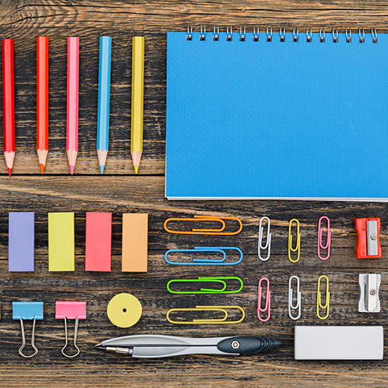 A neatly arranged set of colorful stationery items on a wooden surface exudes an aura of perfectionism, featuring colored pencils, a blue notebook, sticky notes, paper clips, binder clips, a sharpener, an eraser, and a pen.