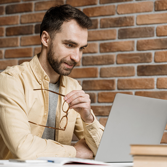 A man with a beard is sitting at a desk, holding glasses in one hand and looking at his laptop screen with meticulous focus. Dressed in a beige shirt over a gray t-shirt, he embodies perfectionism amidst stacks of books against the backdrop of a brick wall.