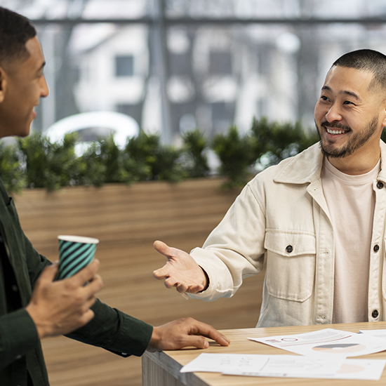 Two men are standing and talking at a table with papers. One holds a coffee cup while the other gestures with mastery. They appear to be in a modern office space with plants and large windows in the background.