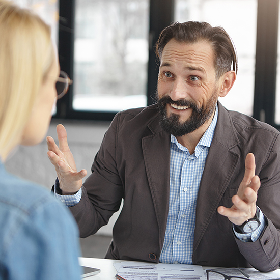 A man with a beard, wearing a blazer and checkered shirt, gestures with mastery as he talks to a woman with long blonde hair, seated across from him. They are at a table with papers in a well-lit room, each focused on the subtle art of persuasion.