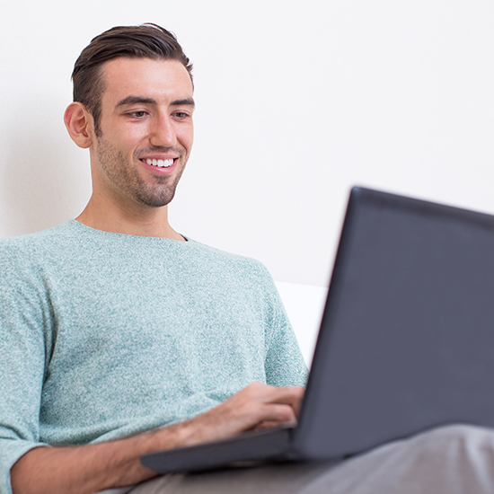 A man sitting comfortably using a laptop, smiling with an air of mastery. He is wearing a light green sweater, and the background is simple and white, creating an almost hypnotic sense of calm.