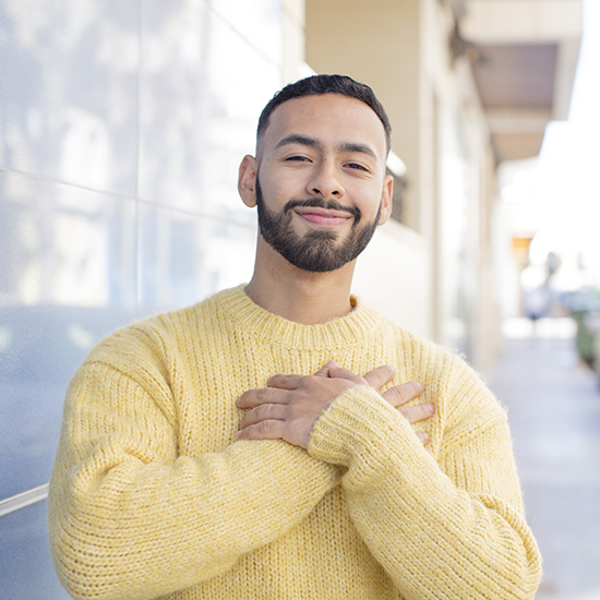 A person with short hair and a beard is smiling and standing outdoors, exuding self-esteem. They are wearing a cozy yellow sweater and have their arms crossed over their chest, appearing content. The background is a blurred urban setting bathed in sunlight.