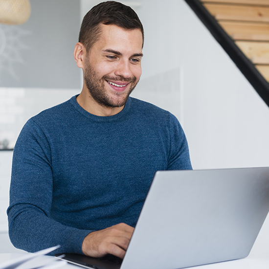 A man in a blue sweater is smiling with self-esteem while working on his laptop. He is seated indoors, with a modern staircase and a light-colored wall in the background.