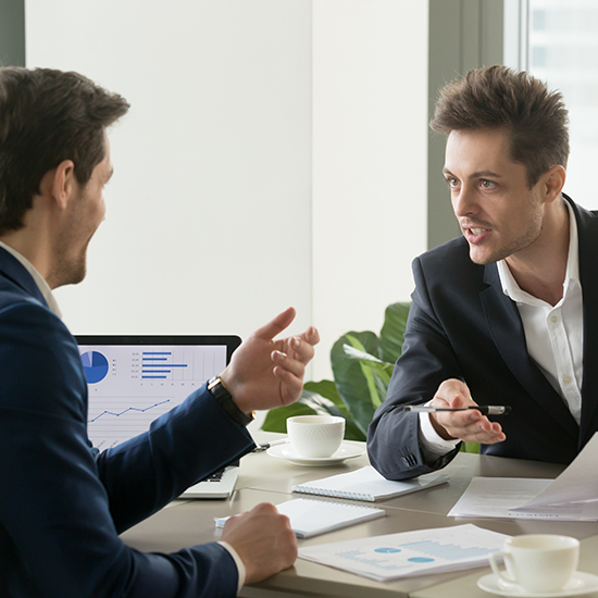 Two men in suits are engaged in a persuasive discussion at a desk with papers and a laptop displaying charts. One man gestures with a pen while they both focus intently. Coffee cups are visible on the desk, enhancing the office's professional atmosphere.