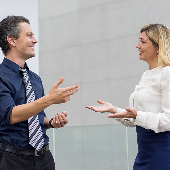 A man and a woman are standing and talking, both smiling and gesturing with their hands. Engaged in a friendly conversation, they seem to be using persuasion techniques in their professional setting, against a neutral background.