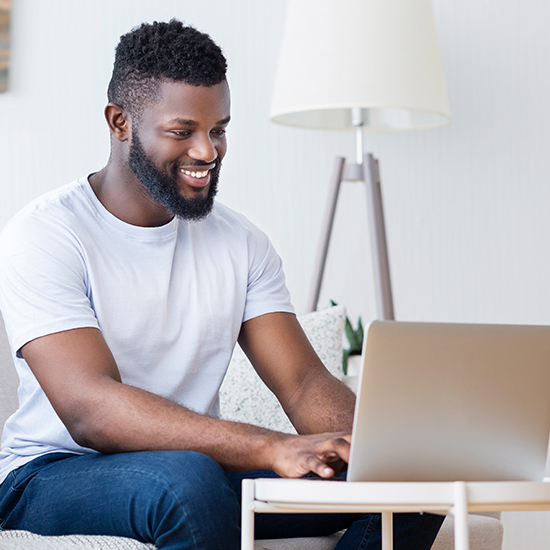 A smiling man with a beard sits on a couch using a laptop, perhaps exploring the art of persuasion. He's wearing a white T-shirt, nestled in a well-lit room with a floor lamp and a light-colored wall in the background.