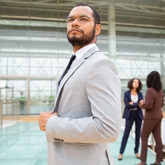 A confident man in a gray suit stands in the foreground, adjusting his tie with courtroom precision. Behind him, two professionals are deep in conversation. The background features a modern glass building interior, reflecting ambition and success.