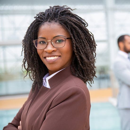 A woman with glasses and braided hair, wearing a brown jacket, smiles confidently with her arms crossed in a brightly lit courtroom. A blurred figure in a suit is visible in the background, adding to the atmosphere of determination and poise.