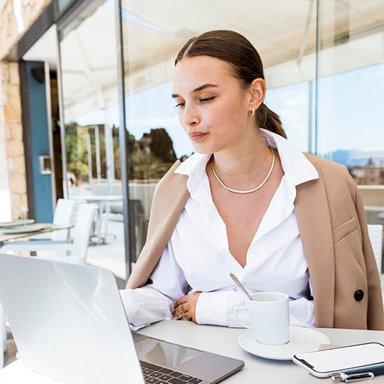 A woman in a white blouse and tan blazer sits confidently at an outdoor café table, focusing on a laptop. A small cup of coffee rests nearby. Glass windows reveal a modern, sunny setting.