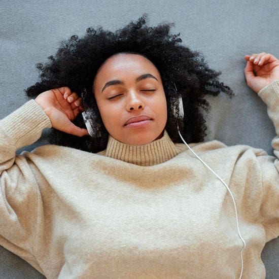 A person with curly hair lies on a gray surface in Orlando, wearing a beige sweater and headphones. Their eyes are closed, appearing relaxed, as if immersed in the soothing rhythms of music or the calming practice of hypnotherapy. A person with curly hair lies on a gray surface in Orlando, wearing a beige sweater and headphones. Their eyes are closed, appearing relaxed, as if immersed in the soothing rhythms of music or the calming practice of hypnotherapy.