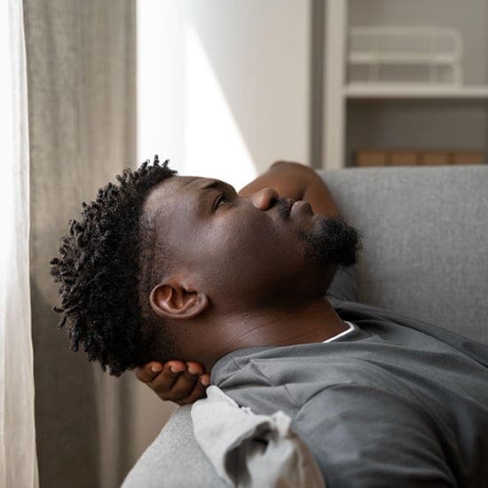 A man with short curly hair lies on a gray sofa in Orlando, looking thoughtfully out a window. He’s wearing a gray T-shirt and has one hand resting under his head. The room is softly lit with natural light, creating a serene atmosphere perfect for hypnotherapy. A man with short curly hair lies on a gray sofa in Orlando, looking thoughtfully out a window. He's wearing a gray T-shirt and has one hand resting under his head. The room is softly lit with natural light, creating a serene atmosphere perfect for hypnotherapy.