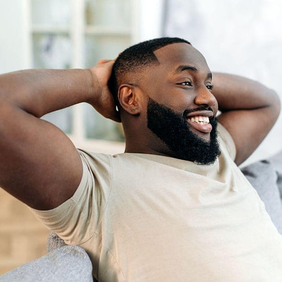 A smiling man with a beard, wearing a light beige t-shirt, relaxes on a couch in a bright room. He exudes the calm demeanor of an Orlando psychologist with his hands casually resting behind his head.