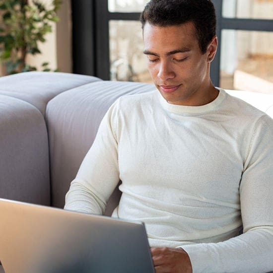 A person in a white sweater sits on a gray couch, typing on a laptop. The background features a large window and some greenery, evoking the cozy ambiance of an Orlando setting, perhaps where a psychologist might unwind after sessions.