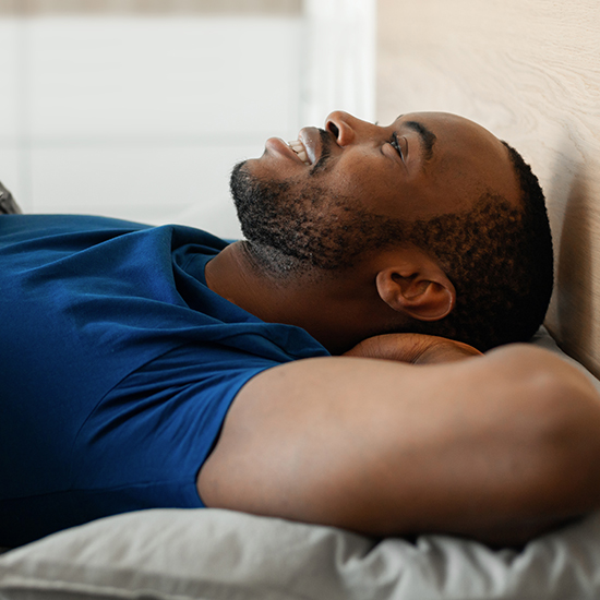 A man with a beard, wearing a blue shirt, is lying on his back, smiling and resting his head on his hands. The background is blurred, emphasizing a peaceful mood as if he’s conquered sleep anxiety and finally found rest.