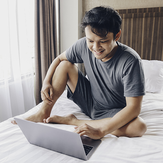 A person sits cross-legged on a bed, smiling and using a laptop. They wear a gray t-shirt and shorts. Sunlight filters through the window with curtains, illuminating the room—a peaceful scene where hypnosis might ease their fear of going to bed.