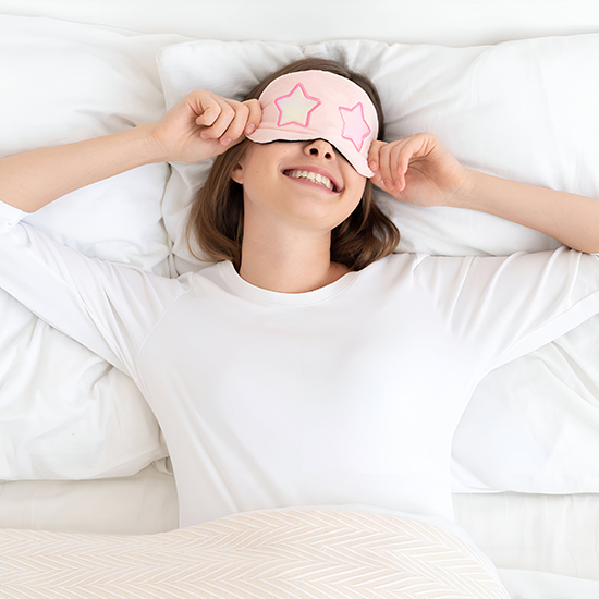 A smiling person lies in bed, wearing a white long-sleeve shirt and a pink sleep mask adorned with star designs, perhaps as part of their new hypnotherapy routine. They gently pull up the mask under a light beige blanket, embracing tranquility and easing their fear of staying awake.