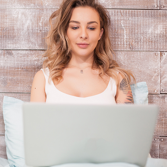 A woman with wavy hair sits against a wooden wall, her laptop resting on her lap. She wears a white sleeveless top, revealing a tattoo on her upper arm as she delves into the screen like someone entranced by hypnosis.