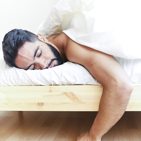 A man with a beard is naturally asleep in bed, lying on his stomach with one arm hanging off the side. The bed, adorned with white sheets, has a wooden frame. The scene exudes peacefulness and calm, almost like a gentle hypnosis.