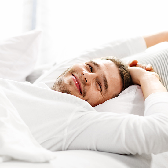 A person with a light beard is lying in bed, smiling while looking at the camera. They are wearing a white shirt, resting their head on a pillow in a bright room, embodying the tranquility often sought through sleep therapy. Their hands are behind their head, suggesting a relaxed and comfortable posture.