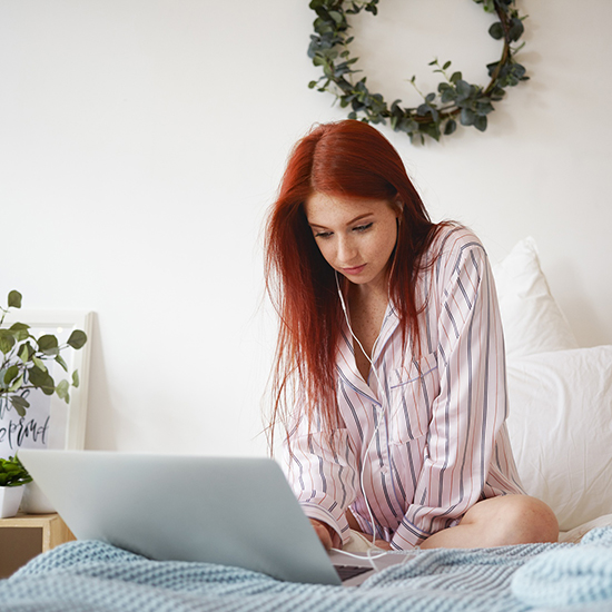 A person with long red hair wearing striped pajamas sits on a bed, using a laptop for hypnotherapy. They have earphones on, perhaps seeking relief from insomnia. The bed features a blue knitted blanket, and a leafy wreath hangs on the white wall beside a nearby plant.