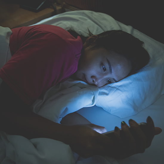 A person lies in bed at night under a white blanket, holding a smartphone with a bright screen illuminating their face. They wear a red shirt and appear engaged with the phone, seemingly caught in the trance of digital hypnosis. The background is dimly lit.