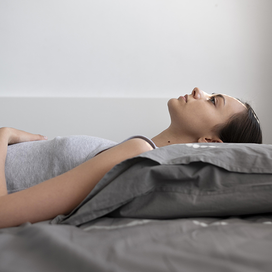 Person lying on a bed, gazing upwards. They are wearing a gray tank top and resting on a gray pillow and bed sheet. The room is softly lit, creating a calm and contemplative atmosphere, reminiscent of sleep hypnosis.